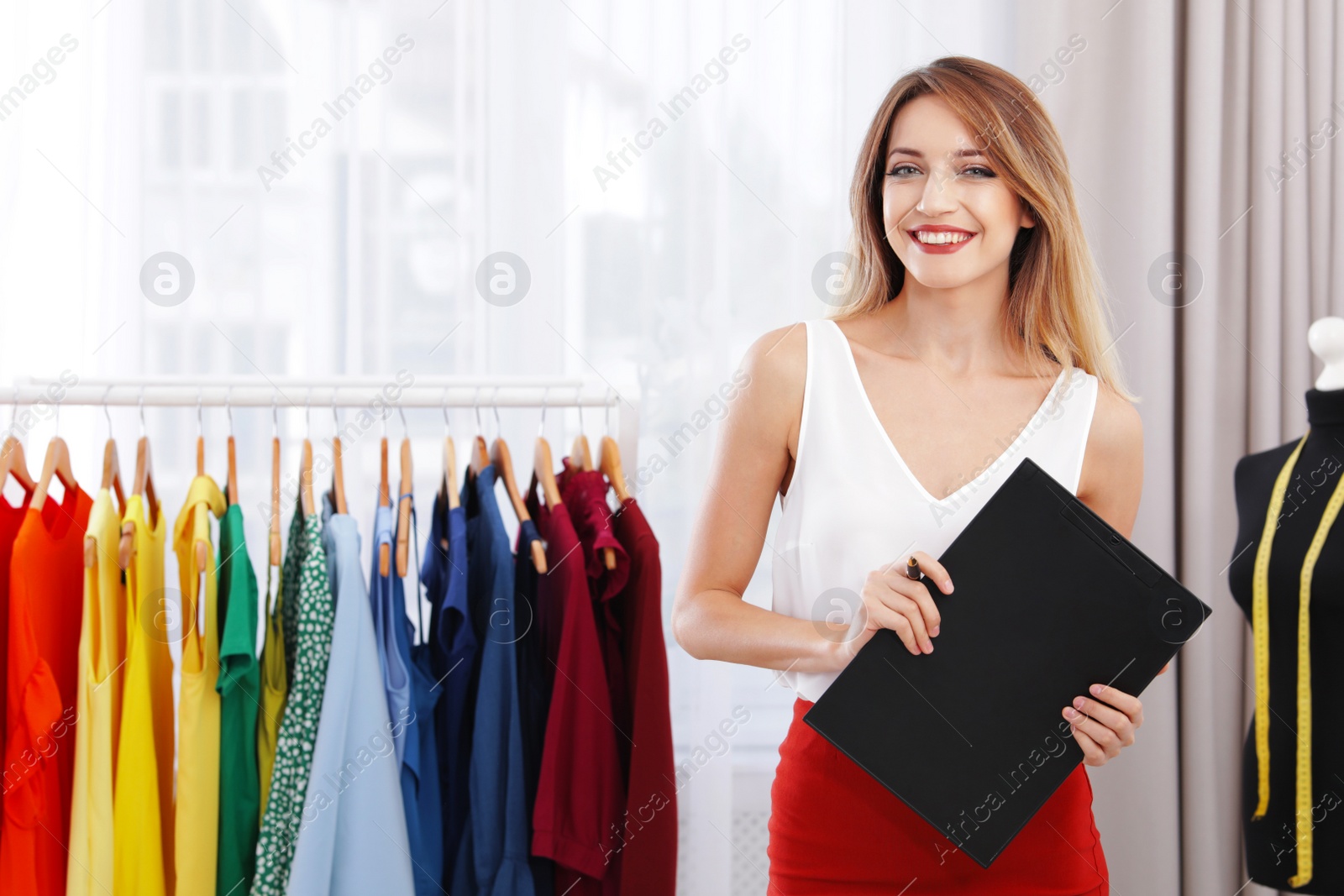 Photo of Beautiful young stylist holding clipboard near rack with designer clothes in studio