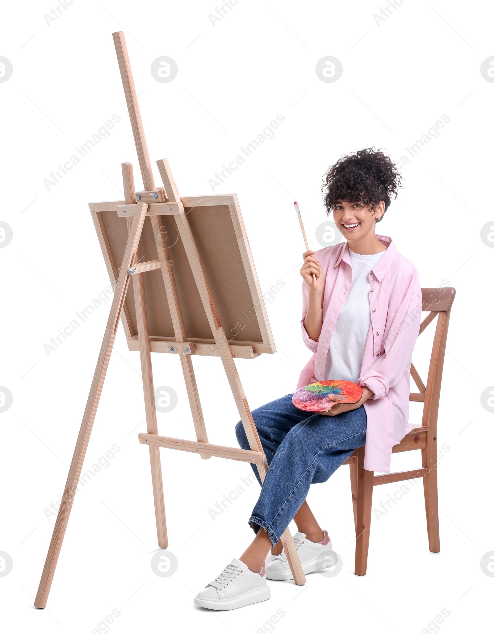 Photo of Young woman holding brush and artist`s palette near easel with canvas against white background