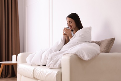 Woman covered in blanket holding cup of drink on sofa