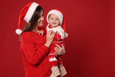 Photo of Happy mother with cute baby in Christmas outfits and Santa hats on red background, space for text