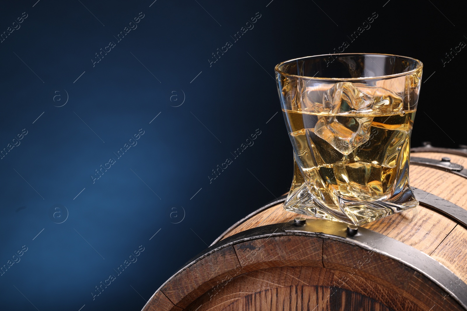 Photo of Whiskey with ice cube in glass on wooden barrel against blue background, space for text