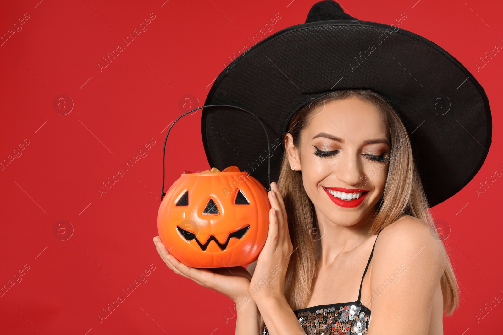 Photo of Beautiful woman wearing witch costume with Jack O'Lantern candy container on red background. Halloween party