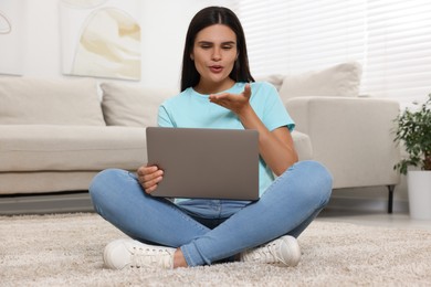 Photo of Young woman having video chat via laptop and blowing kiss on floor at home