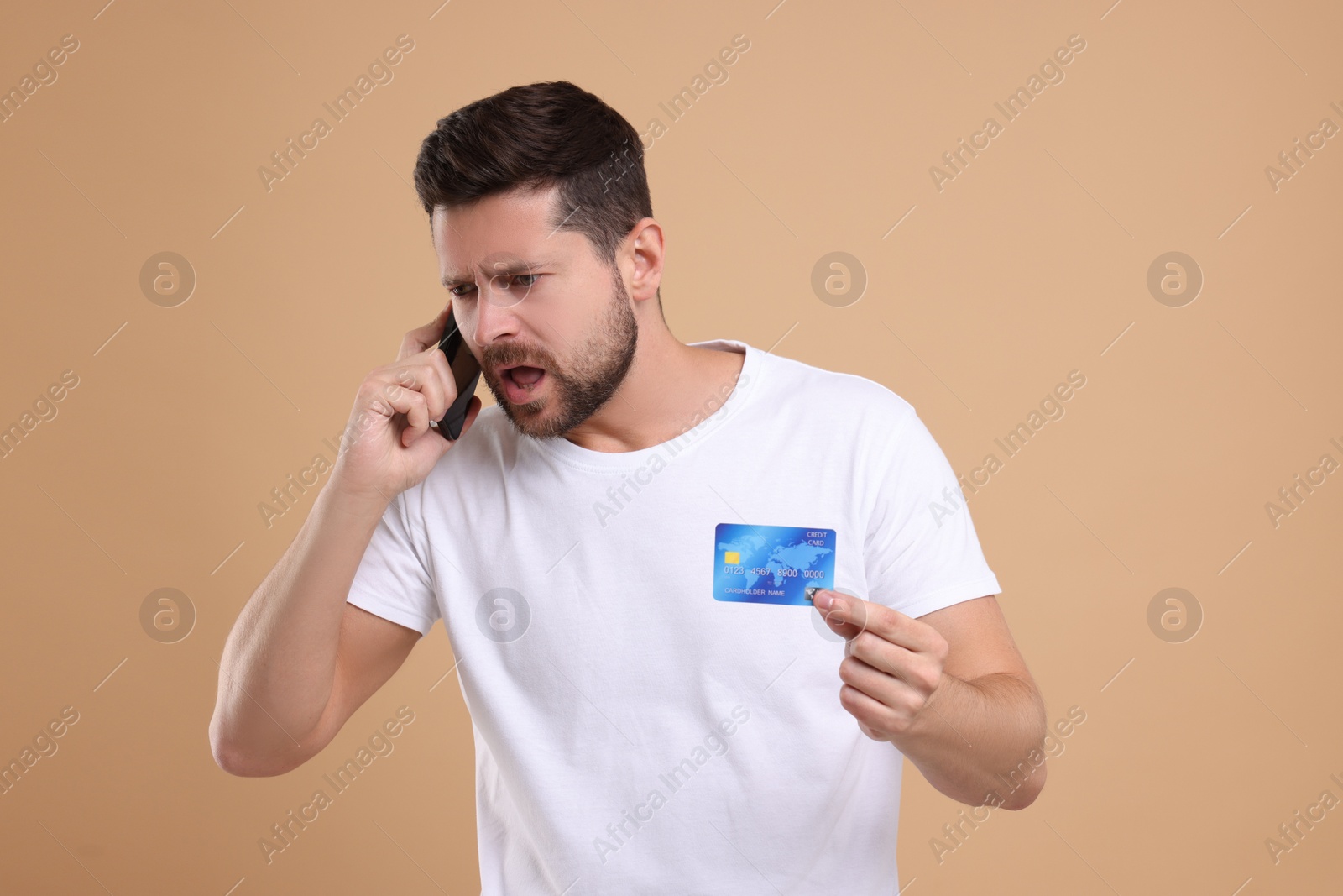 Photo of Emotional man with credit card talking on smartphone against beige background. Be careful - fraud