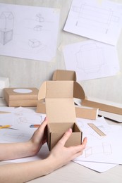 Photo of Woman holding cardboard box near drawings at table, closeup