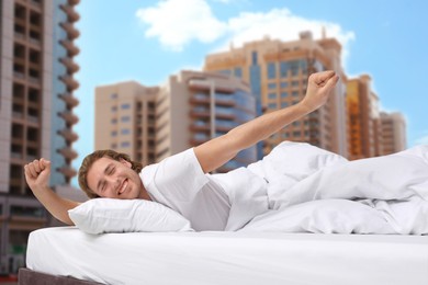 Image of Happy man stretching in bed and beautiful view of cityscape on background. Good sleep despite of urban noise