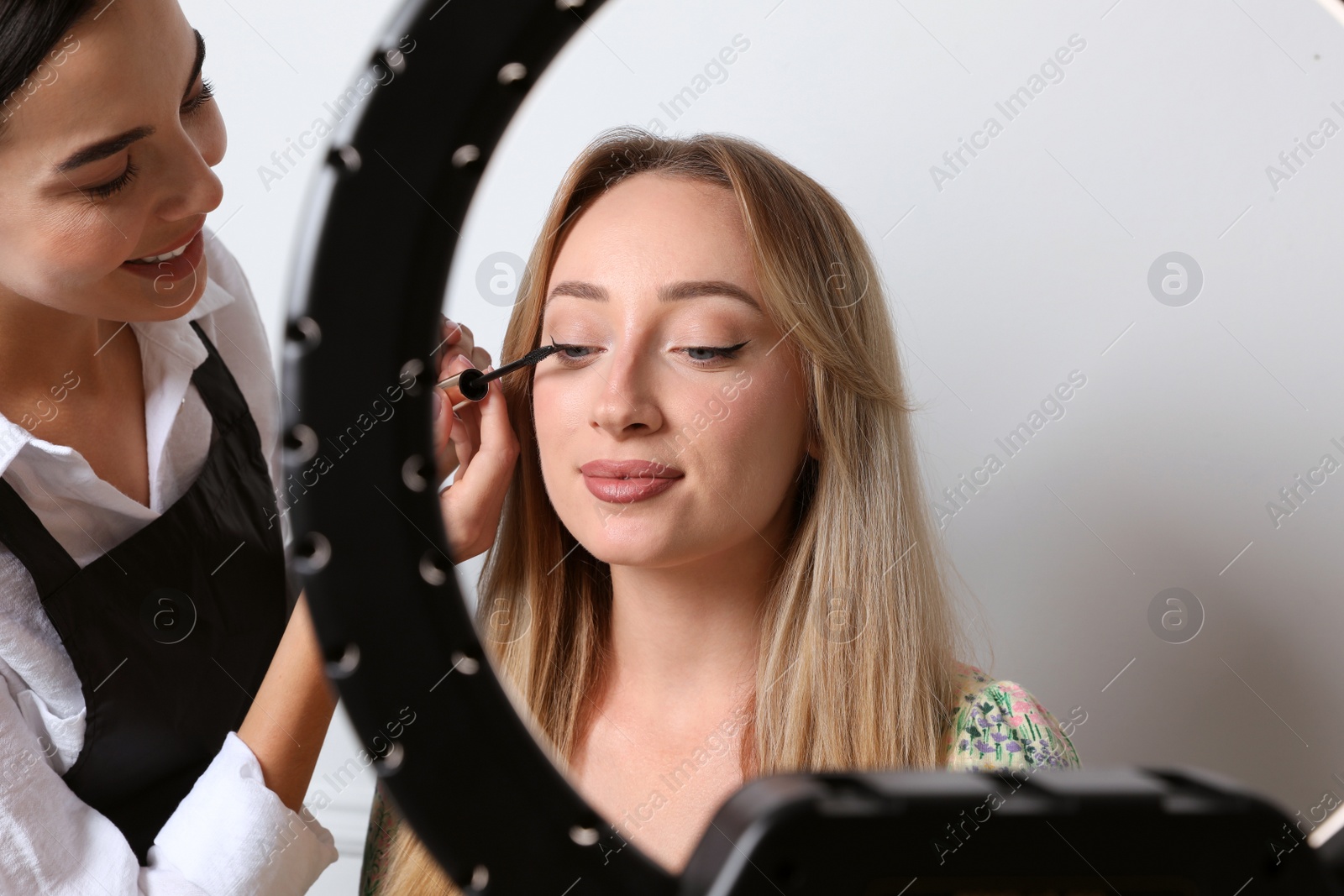 Photo of Professional makeup artist working with beautiful young woman against light background, view through ring lamp