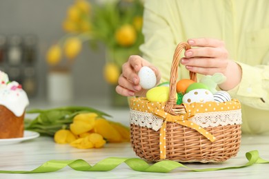 Photo of Closeup of woman putting painted egg into Easter basket at white marble table. Space for text