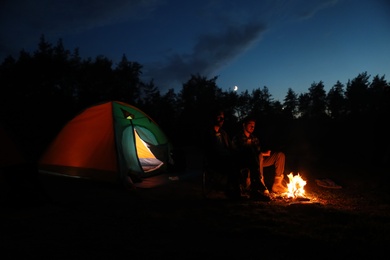 Young men sitting near bonfire and camping tent in wilderness at night