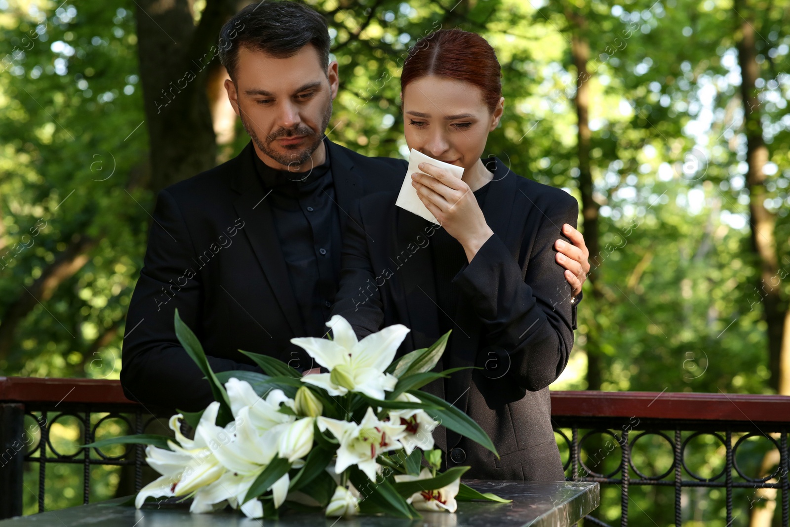 Photo of Sad couple mourning near granite tombstone with white lilies at cemetery. Funeral ceremony