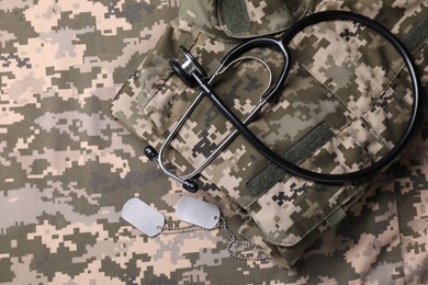 Photo of Stethoscope, military ID tags and soldier uniform on camouflage fabric, flat lay