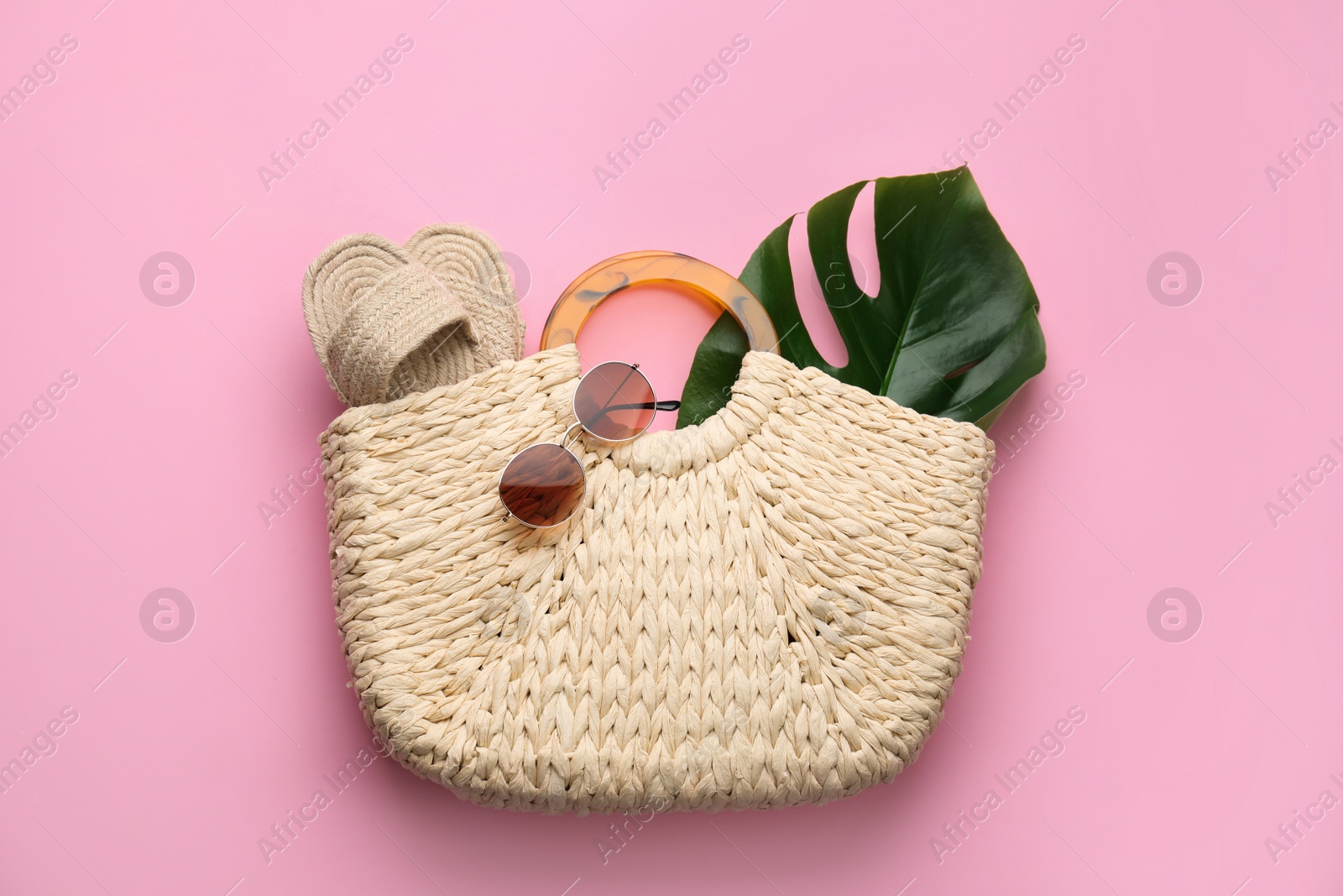 Photo of Elegant woman's straw bag with shoes, tropical leaf and sunglasses on pink background, top view