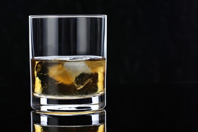 Photo of Tasty whiskey and ice cubes in glass on mirror table against black background, closeup. Space for text