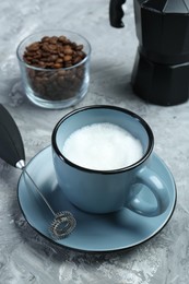 Photo of Mini mixer (milk frother), whipped milk in cup and coffee beans on grey textured table