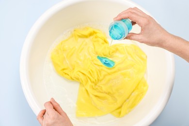 Woman pouring detergent into basin with baby clothes on light blue background, top view