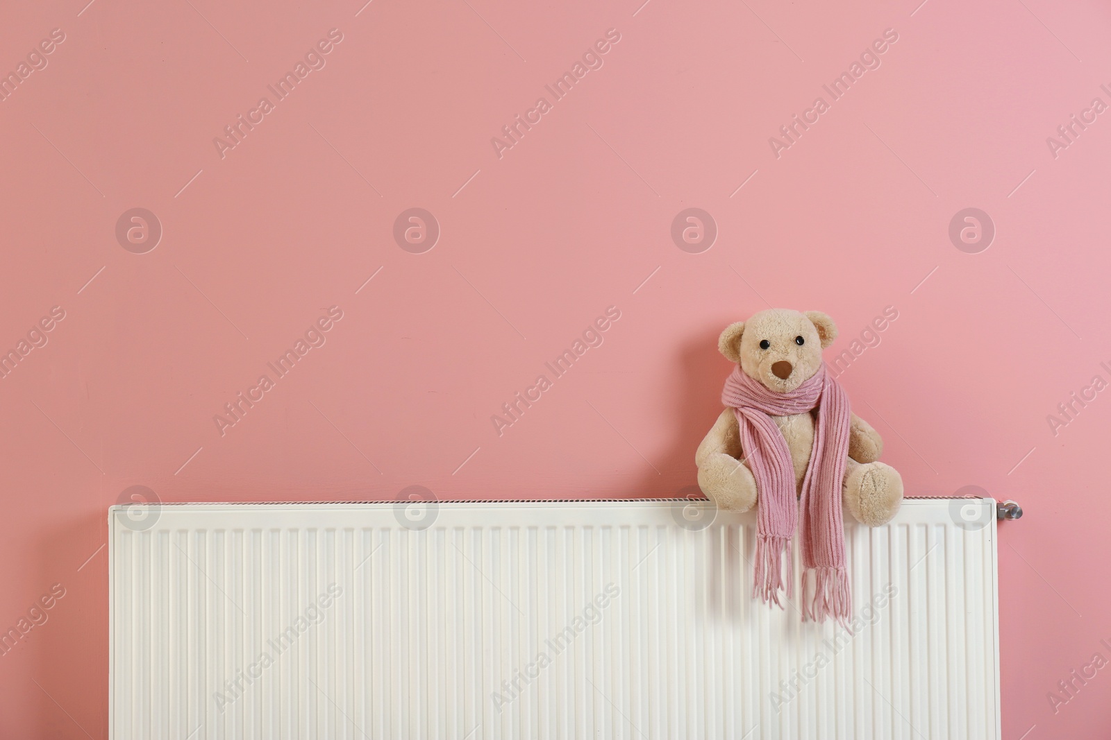 Photo of Teddy bear with knitted scarf on heating radiator near color wall. Space for text