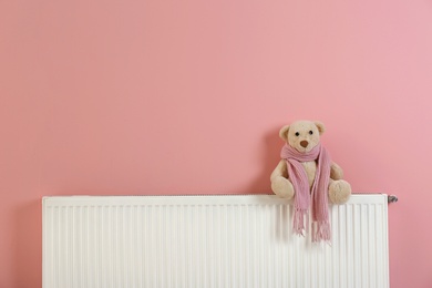 Teddy bear with knitted scarf on heating radiator near color wall. Space for text