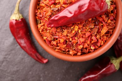 Photo of Chili pepper flakes in bowl and pods on dark textured table, flat lay