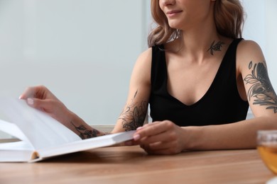 Photo of Beautiful woman with tattoos on body reading book at table indoors, closeup