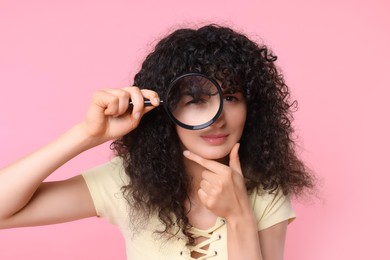 Curious young woman looking through magnifier glass on pink background
