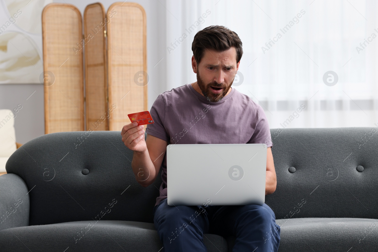 Photo of Emotional man with laptop and credit card on sofa at home. Be careful - fraud