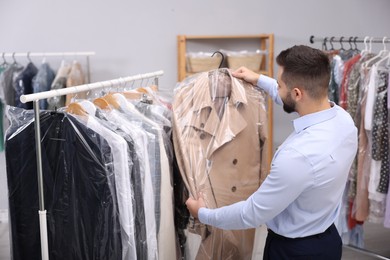 Photo of Dry-cleaning service. Worker holding hanger with coat in plastic bag near rack with clothes indoors