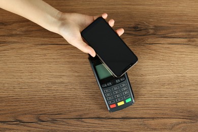 Woman with smartphone using modern payment terminal at wooden table, top view