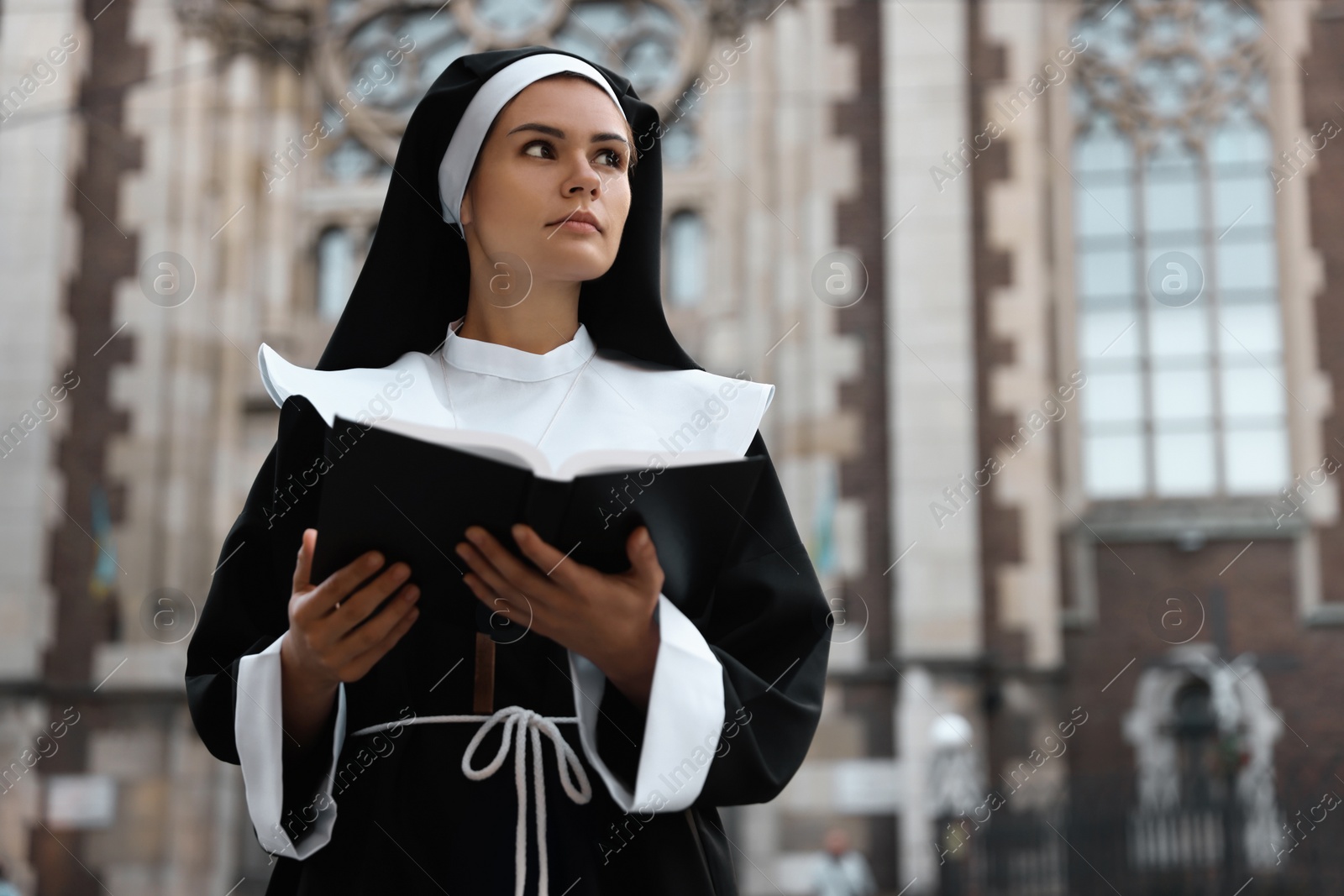 Photo of Young nun reading Bible near cathedral outdoors. Space for text