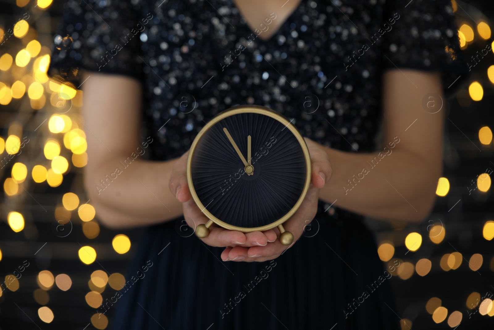 Photo of Woman holding alarm clock against blurred lights, closeup. New Year countdown