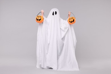Photo of Child in white ghost costume holding pumpkin buckets on light grey background. Halloween celebration