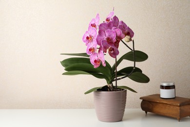 Photo of Beautiful blooming orchid, jar and old wooden jewelry box on white table near beige wall. Space for text