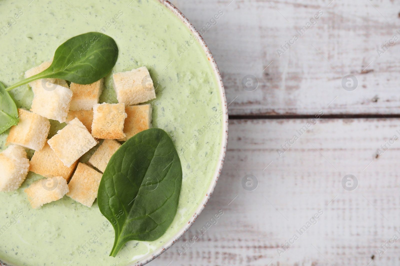 Photo of Delicious spinach cream soup with leaves and croutons in bowl on white wooden table, top view. Space for text