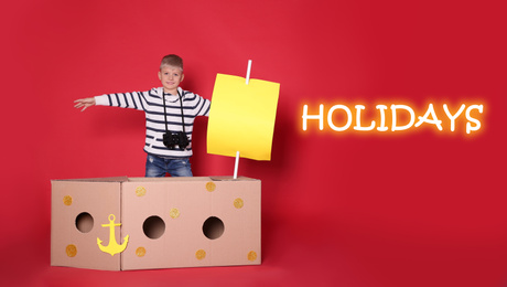 School holidays. Cute little child playing with cardboard ship near red wall. Banner design 
