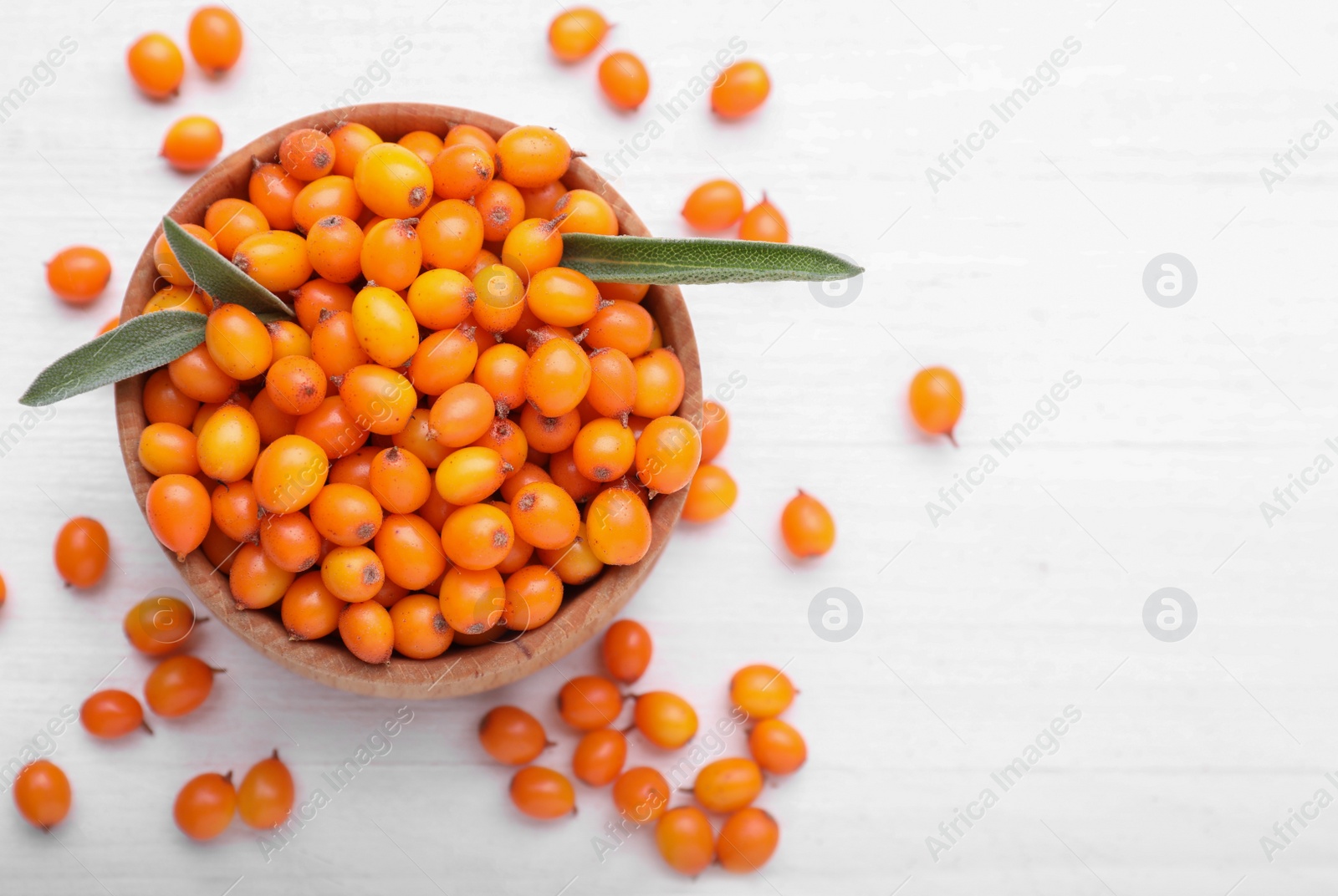 Photo of Bowl with fresh ripe sea buckthorn berries and green leaves on white wooden table, flat lay. Space for text