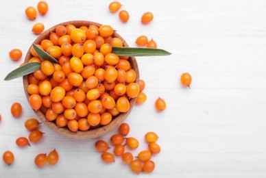 Photo of Bowl with fresh ripe sea buckthorn berries and green leaves on white wooden table, flat lay. Space for text