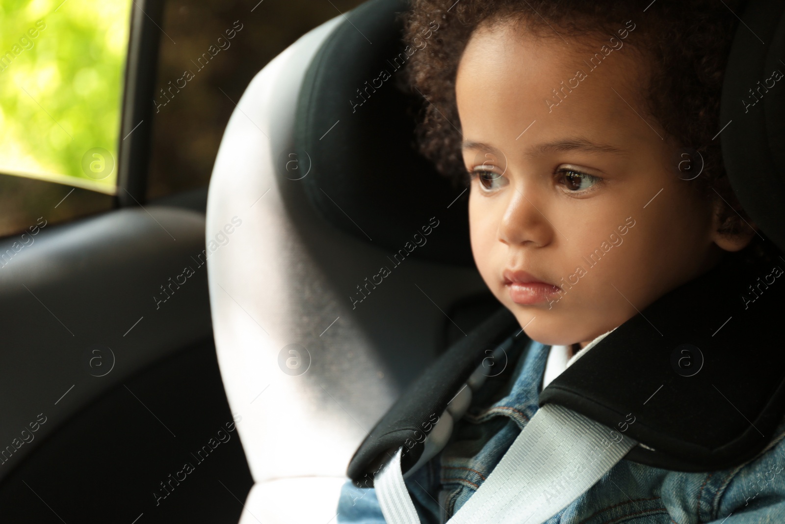 Photo of Cute African-American girl sitting in safety seat alone inside car. Child in danger