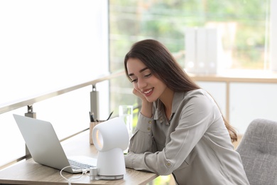 Young woman enjoying air flow from portable fan at workplace. Summer heat