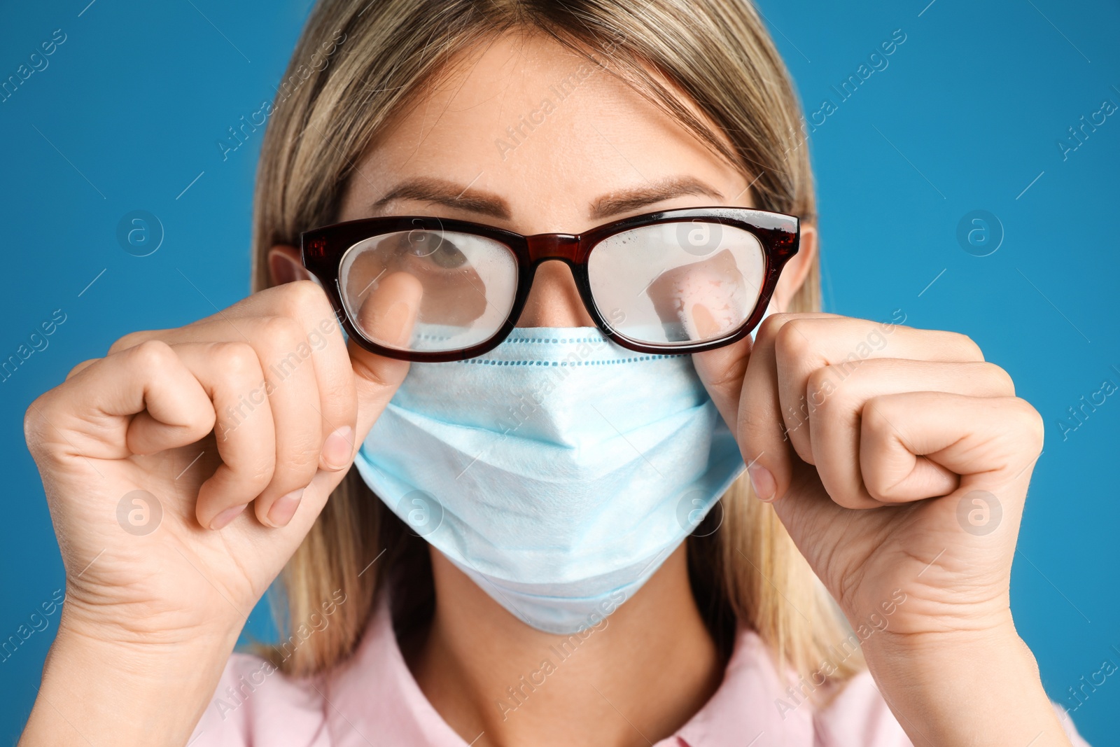 Photo of Woman wiping foggy glasses caused by wearing disposable mask on blue background, closeup. Protective measure during coronavirus pandemic