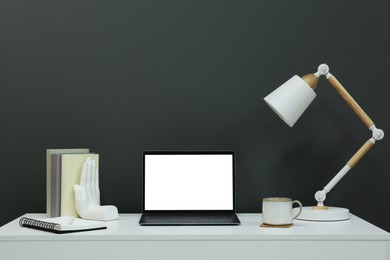 Stylish workplace with laptop, lamp, cup and decor on white table near grey wall