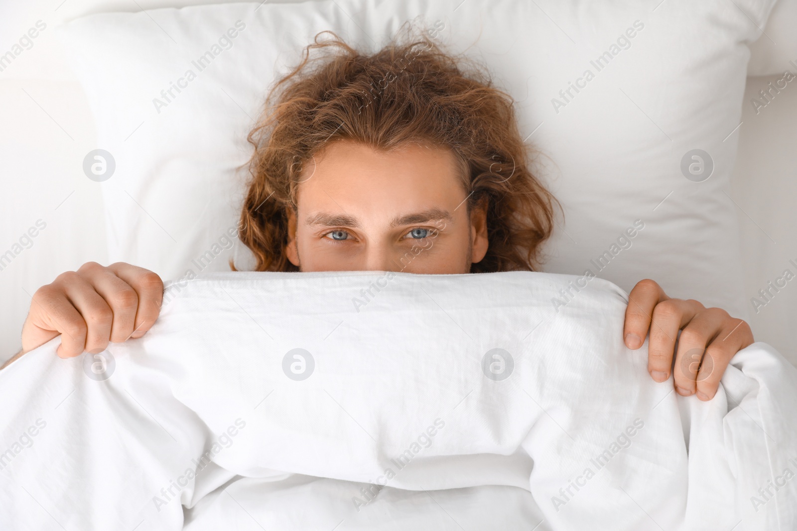 Photo of Young man covering his face with blanket while lying on pillow, top view. Bedtime