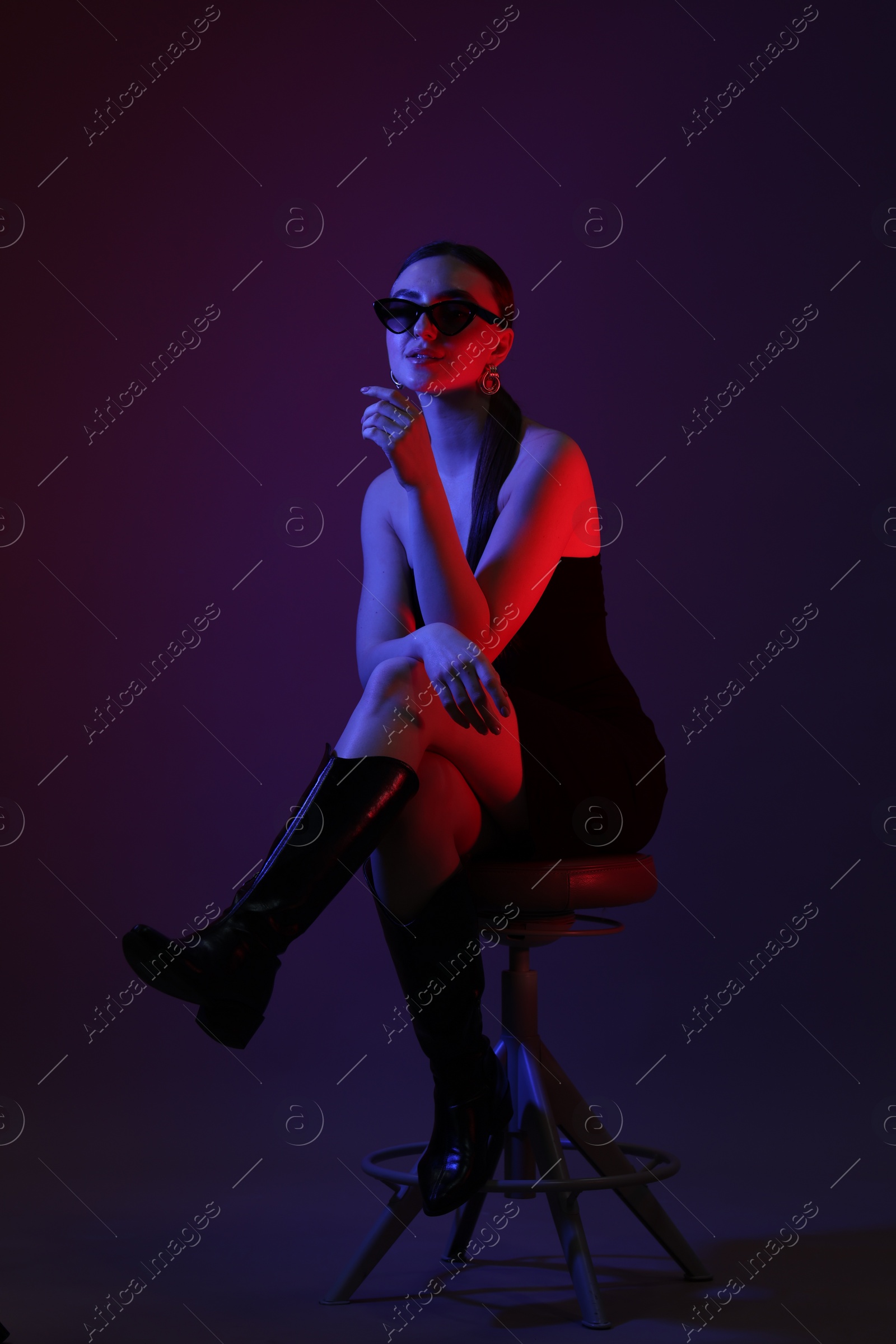 Photo of Beautiful young woman in sunglasses posing on stool against color background with neon lights