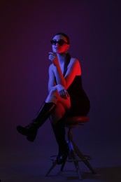 Photo of Beautiful young woman in sunglasses posing on stool against color background with neon lights