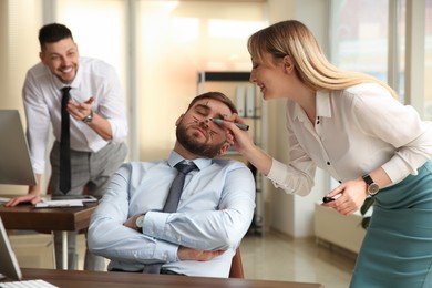 Photo of Young woman drawing on colleague's face while he sleeping in office. Funny joke