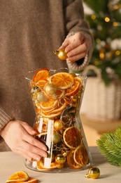 Photo of Woman putting Christmas tree ball into glass vase with dry orange slices and cones at white wooden table, closeup, Festive decor