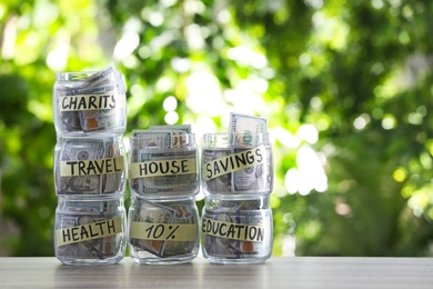 Glass jars with money for different needs on table against blurred background