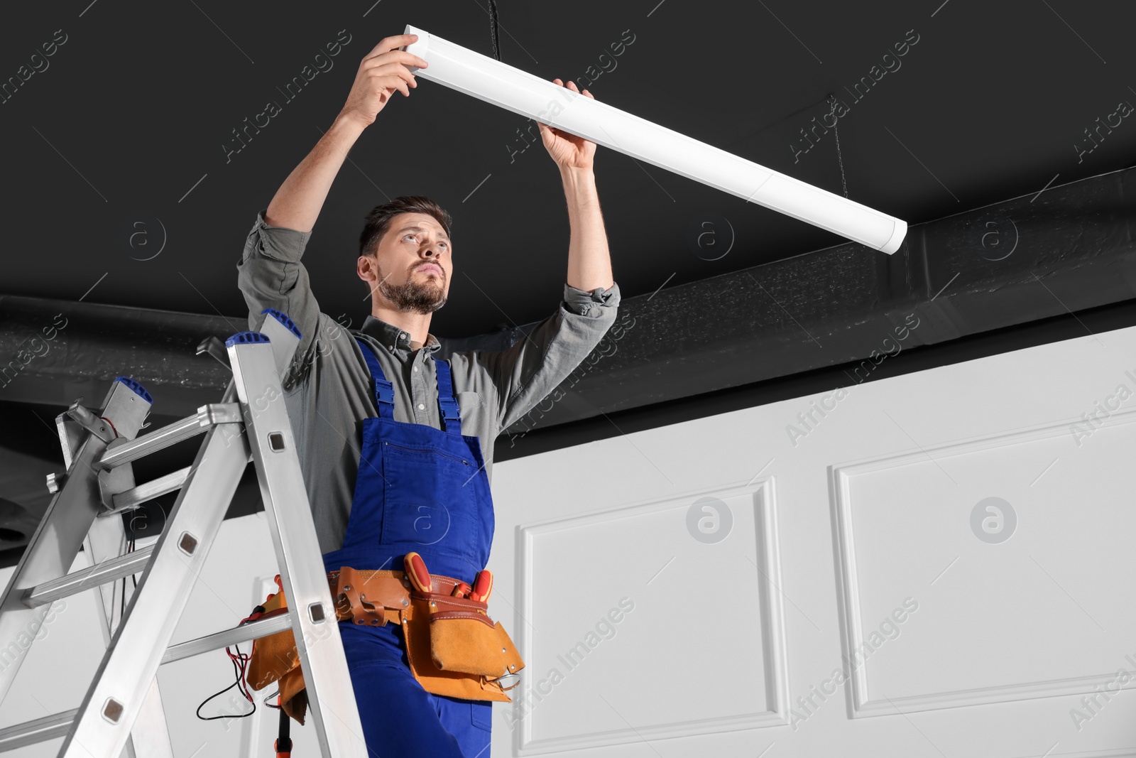 Photo of Electrician in uniform installing ceiling lamp indoors