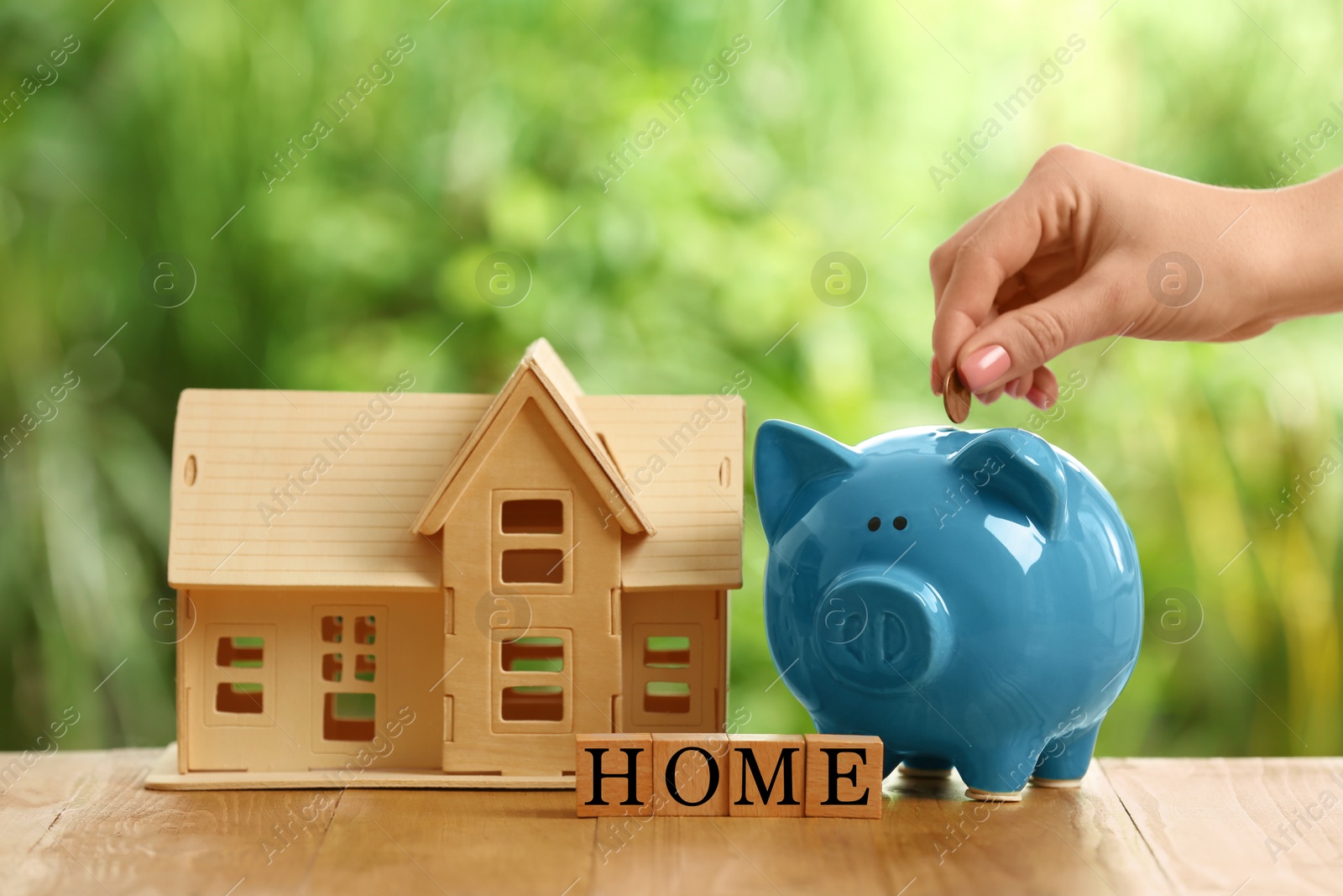 Photo of Woman putting money into piggy bank, house model and word Home made of cubes on wooden table outdoors, closeup