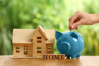 Photo of Woman putting money into piggy bank, house model and word Home made of cubes on wooden table outdoors, closeup