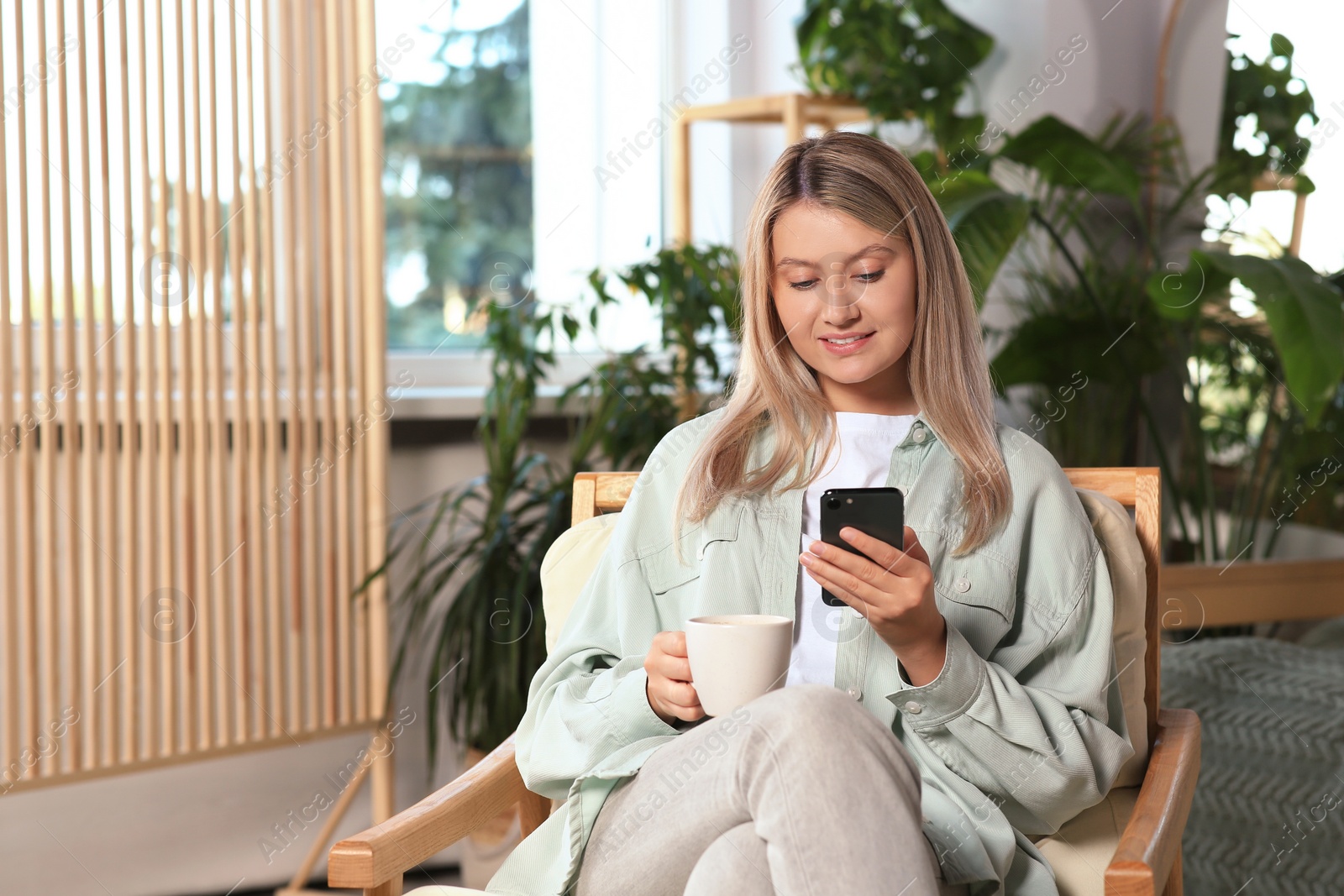 Photo of Woman with smartphone and cup of drink sitting in armchair at home