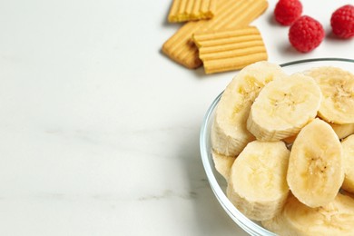 Photo of Cut bananas in glass bowl and other baby finger foods on white marble table, space for text